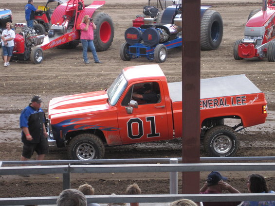 Orange pickup truck with "General Lee" and "01" on the side, in a dirt arena with other vehicles