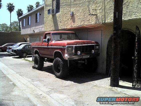 Red 1978 Ford Bronco pickup truck with lifted suspension and large tires parked in front of a building