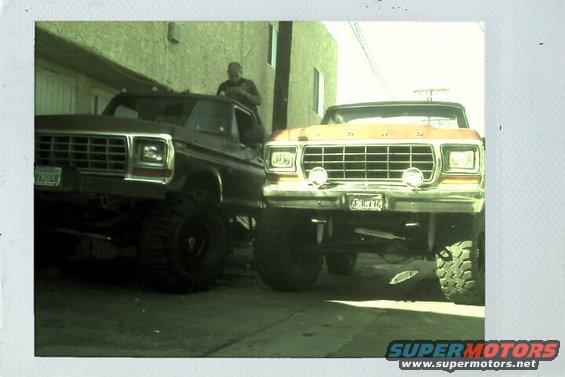 Two lifted Ford trucks parked side-by-side, front view. The truck on the right is orange