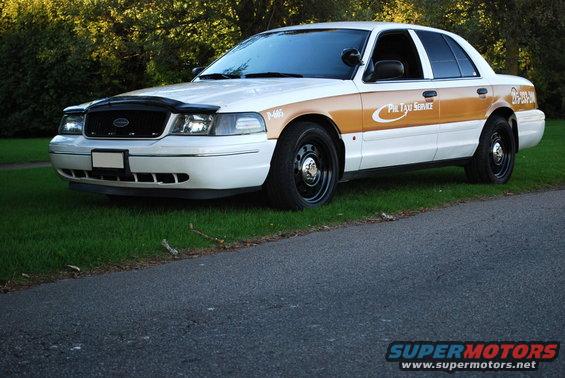 White and tan 1998 Ford Crown Victoria taxi with black wheels, parked on grass next to a road