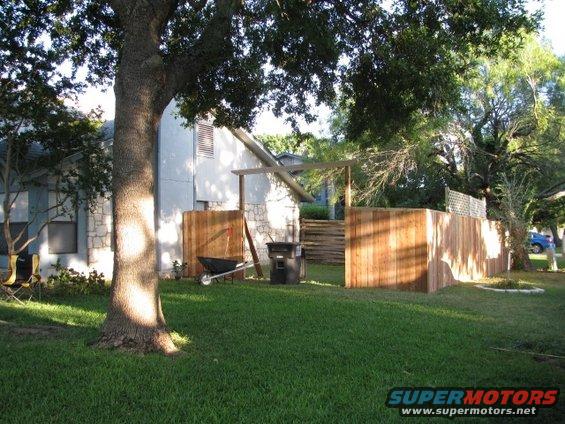 Backyard with a large tree, grass, wooden fence, and a blue vehicle in the distance