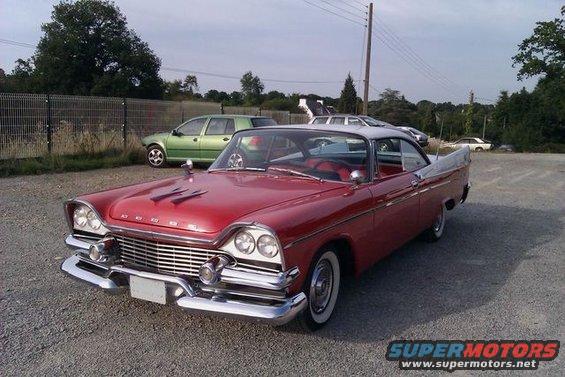 Red and white classic car with chrome grille and fins, parked on gravel lot