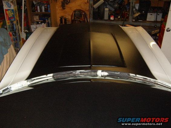 White and black car hood with a center bulge, showing windshield wipers and a portion of the windshield trim