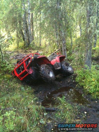 Two red ATVs are stuck in a muddy trail in a wooded area