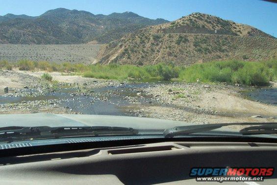 View from inside a vehicle looking out at a dry riverbed and mountains. Supermotors.net logo visible