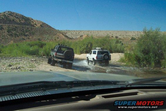 Two SUVs driving through a riverbed with scrub brush and hills in the background
