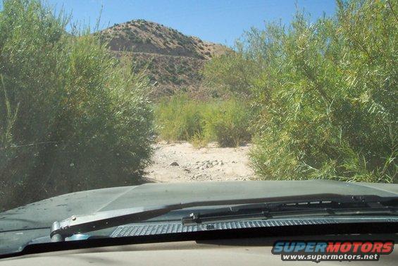 Dashboard view from a vehicle driving through a dry creek bed with brush and a hill in the background. The windshield wipers are visible