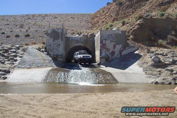 White SUV exiting a tunnel and driving through water. It has a roof rack