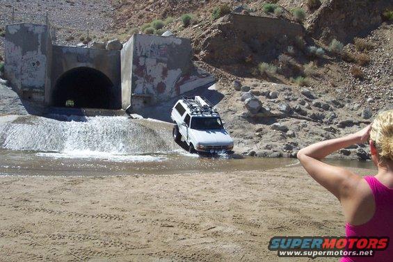 White SUV with roof rack driving through shallow water near a concrete tunnel