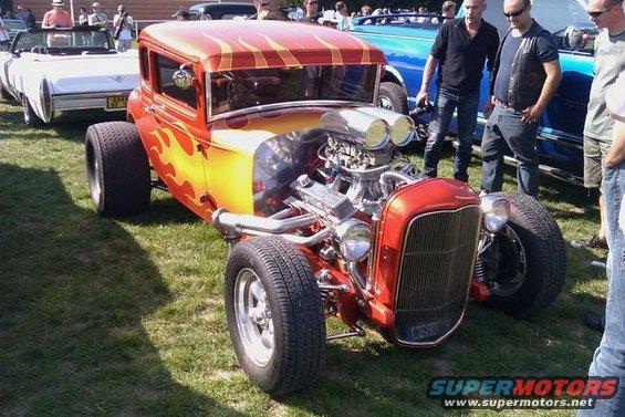 Red hot rod with flames, dual carburetors, and exposed engine. Other cars and people in background
