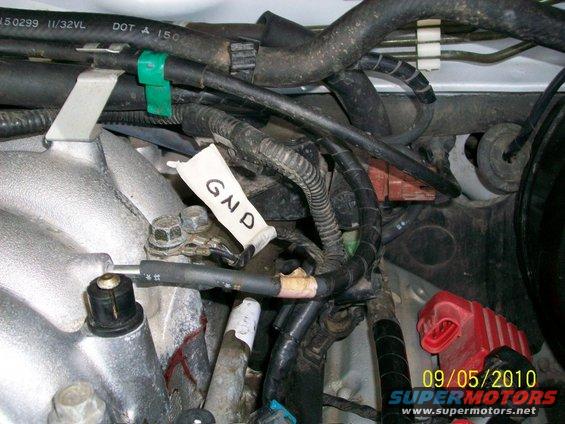 Engine bay with wires, hoses, and a white tag labeled GND. A red battery terminal cover is visible