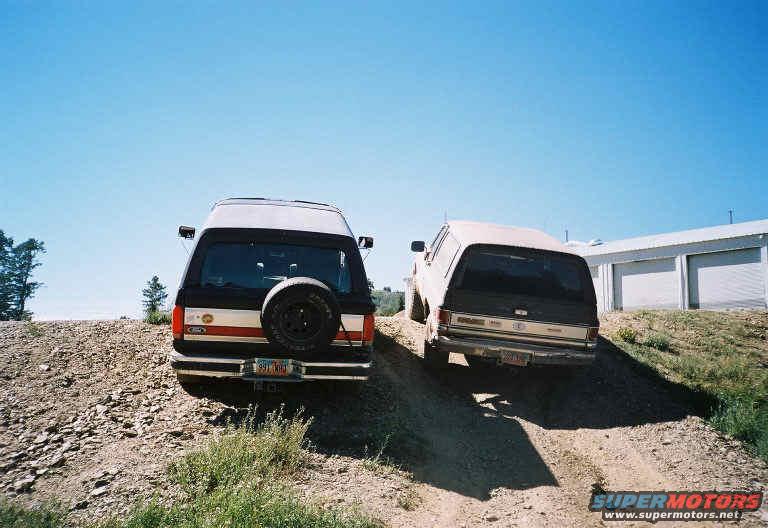 Rear view of a black and red 1992 Ford Bronco with a spare tire mounted on the back, parked on a dirt hill next to another SUV