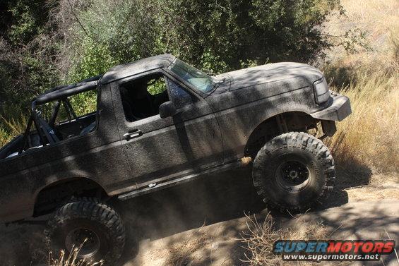 1992 Ford Bronco covered in mud, driving on a dirt trail with large off-road tires