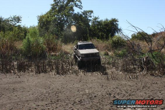 Pickup truck stuck in muddy terrain with dry brush and trees in background