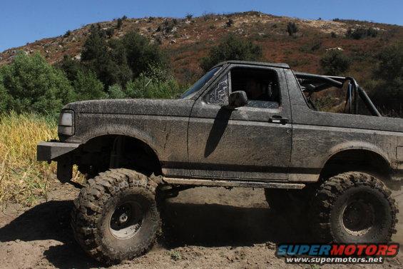 Muddy 1992 Ford Bronco with large tires on a dirt trail, hills in background