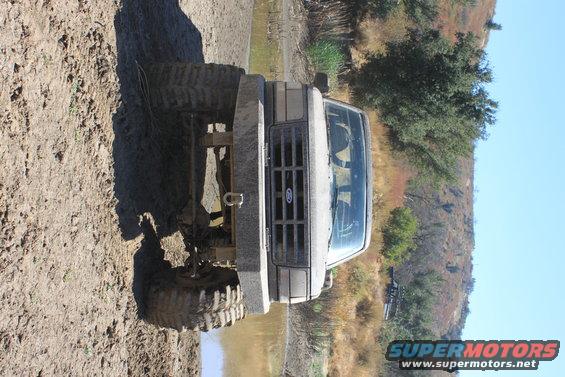 Front view of a lifted, muddy truck with large tires, parked on dirt and in shallow water. Hills and trees in background