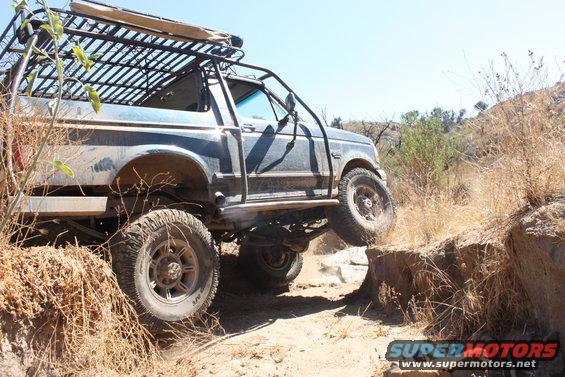 Ford Bronco with a roof rack and roll cage driving on a dirt trail, tires on rocks
