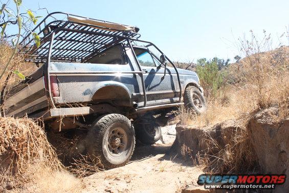 Blue 1992 Ford Bronco with a roof rack and roll cage, off-roading on a dirt trail