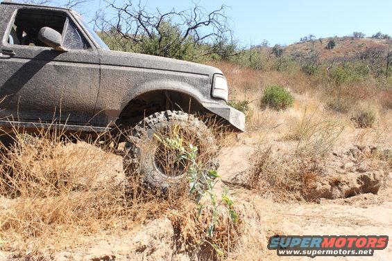 Mud-caked SUV driving off-road, front wheel visible, dry brush and hills in background