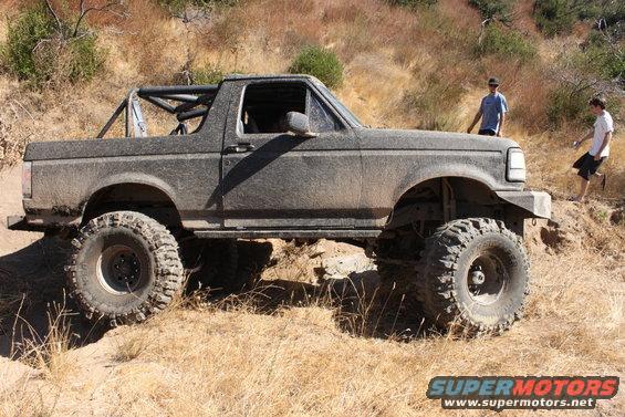 Mud-covered 1992 Ford Bronco with roll cage and large off-road tires on a dirt trail