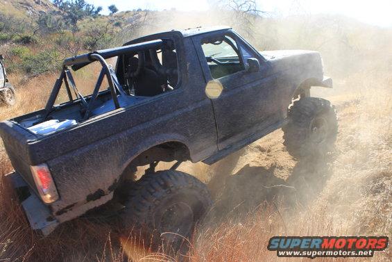 Dark 1992 Ford Bronco off-roading, kicking up dust, with a roll cage and large tires