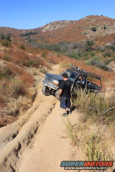 Black 1992 Ford Bronco off-roading on a dirt trail, with a man standing beside it