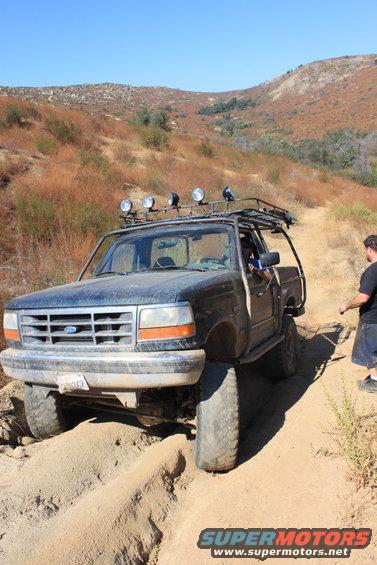 1992 Ford Bronco with roof rack and off-road lights navigating a sandy trail with a person standing nearby