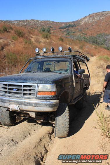 Dark blue Ford Bronco with a roof rack and off-road lights driving on a dirt trail, getting stuck in a rut. A man stands nearby
