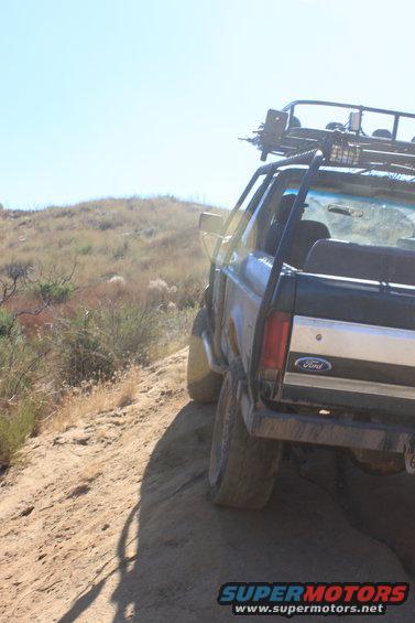 Rear view of a Ford Bronco with a roof rack driving on a dirt trail