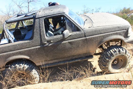Mud-covered 1992 Ford Bronco with roll cage and large off-road tires on a dirt trail