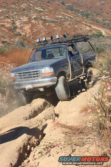 Ford Bronco navigating a rutted dirt trail with off-road lights on the roof rack