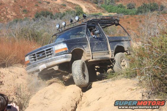 1992 Ford Bronco climbing a steep dirt hill with off-road lights and roof rack