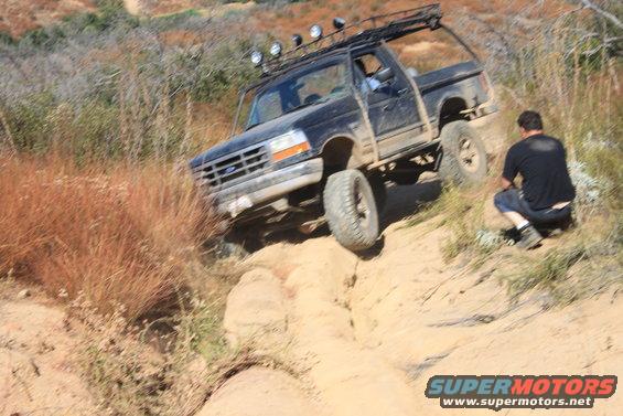 Black 1992 Ford Bronco with roof rack and off-road tires climbing a sandy hill