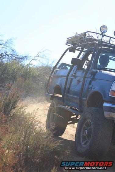 1992 Ford Bronco with roll cage and roof rack driving on a dusty trail