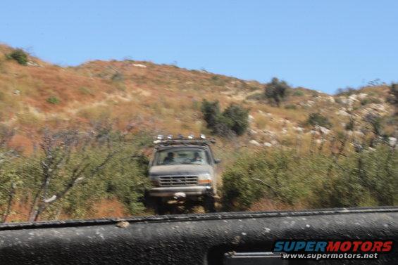 1992 Ford Bronco driving on a dirt trail with brush and hills in the background