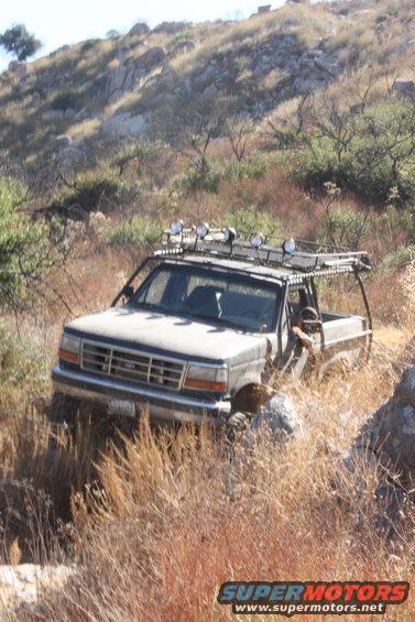 Gray 1992 Ford Bronco with a roof rack and off-road lights, partially obscured by dry brush on a rocky hillside