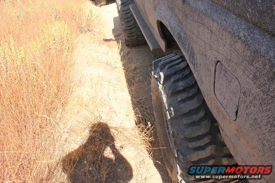 Off-road tire and fender of a vehicle, with dry brush and a shadow in the foreground. Supermotors.net logo visible