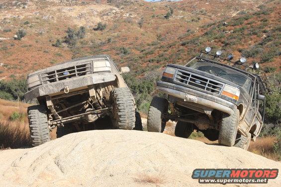 Two lifted Ford Broncos climbing a rocky hill off-road. One is tan, the other is black with extra lights
