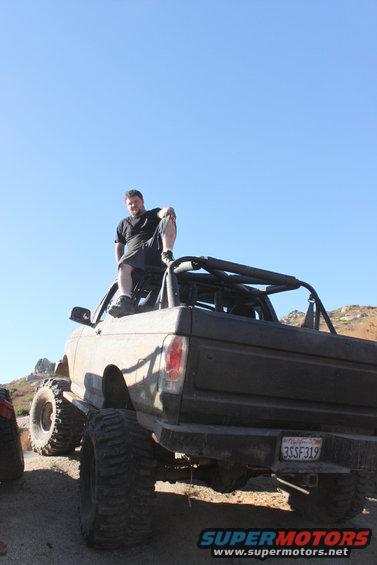 Lifted off-road truck with large tires, roll cage, and a man sitting on top. The vehicle is dusty and appears to be in a desert environment