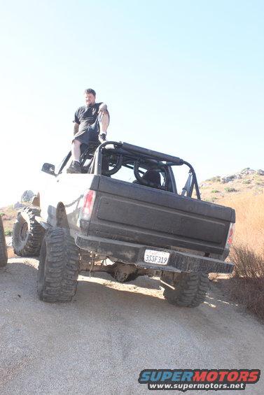 Rear view of a lifted, modified 1980s pickup truck with large tires and a roll cage. A man sits on the roof
