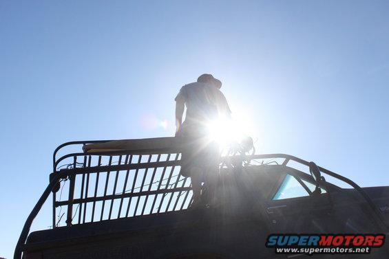 Person standing on the roof rack of a dark vehicle, silhouetted against the sun