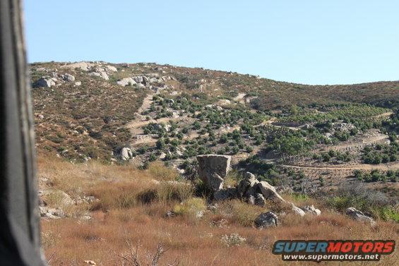 Dry grass and rocks on a hillside with scattered trees and a clear blue sky. Supermotors logo in the corner