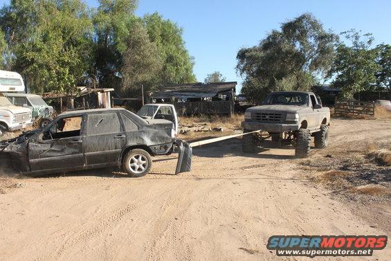 Damaged black sedan towed by a lifted gray pickup truck in a junkyard