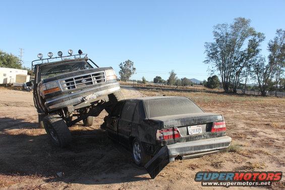 Lifted Ford truck on top of a dark sedan, crushing it. Truck has lights on roof. Sedan has license plate visible