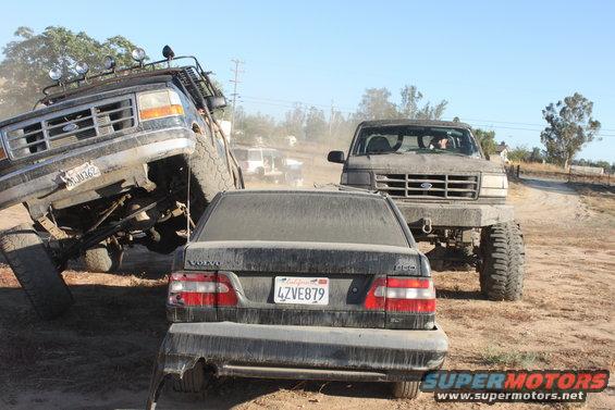 Two lifted Ford trucks are off-roading, one is climbing over a Volvo sedan