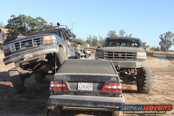 Two lifted Ford trucks, one on its side, with a Volvo sedan in front. License plate 4ZV E879 visible