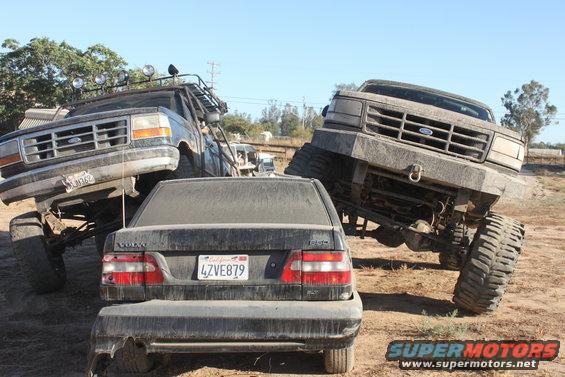 Two lifted Ford trucks tower over a Volvo sedan in a dusty outdoor scene