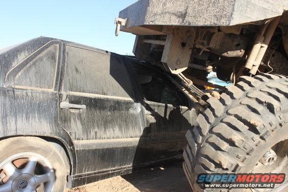 Black sedan with a crushed side, partially obscured by a large off-road vehicle's tire and chassis