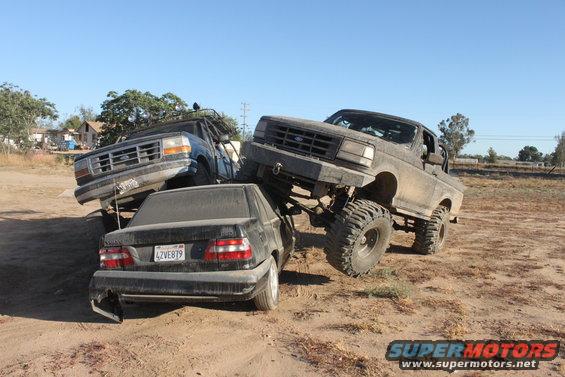 Two lifted Ford trucks on a dirt lot, one crushing a sedan