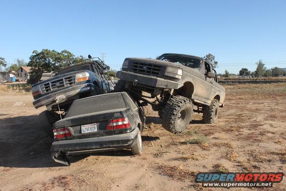 Two lifted Ford trucks are crushing a black sedan in a dirt lot. The front truck is a dark color, the rear truck is blue. The sedan's license plate is visible
